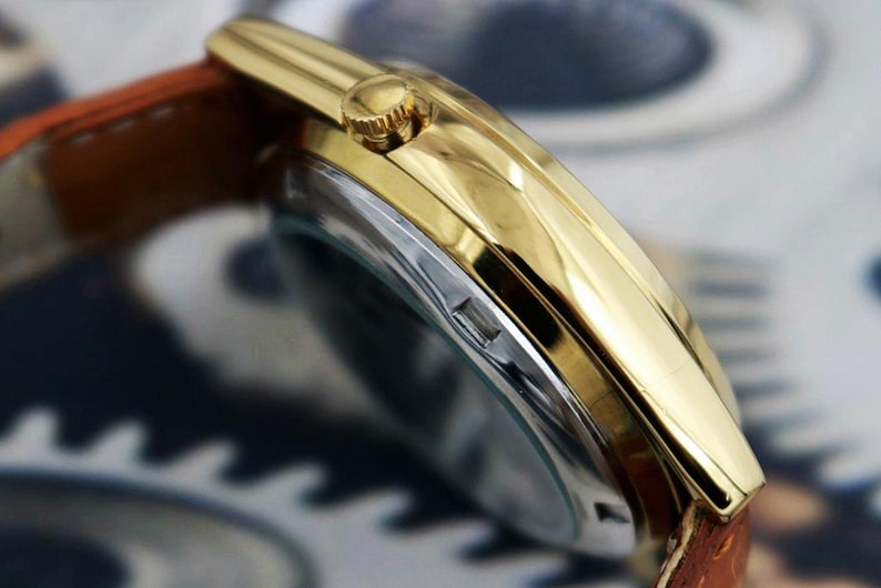 May include: Close-up of a gold-toned wristwatch with a brown leather band. The watch has a gold bezel and a silver-colored face. The band is a light brown color.