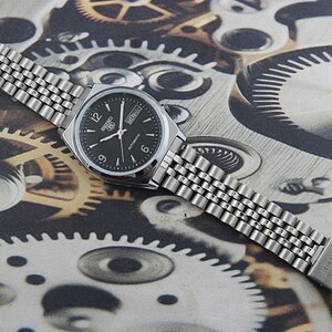 May include: A silver-toned Seiko automatic wristwatch with a black dial, featuring white hour markers and hands. The watch has a stainless steel band and is displayed on a background of gears and mechanical components.