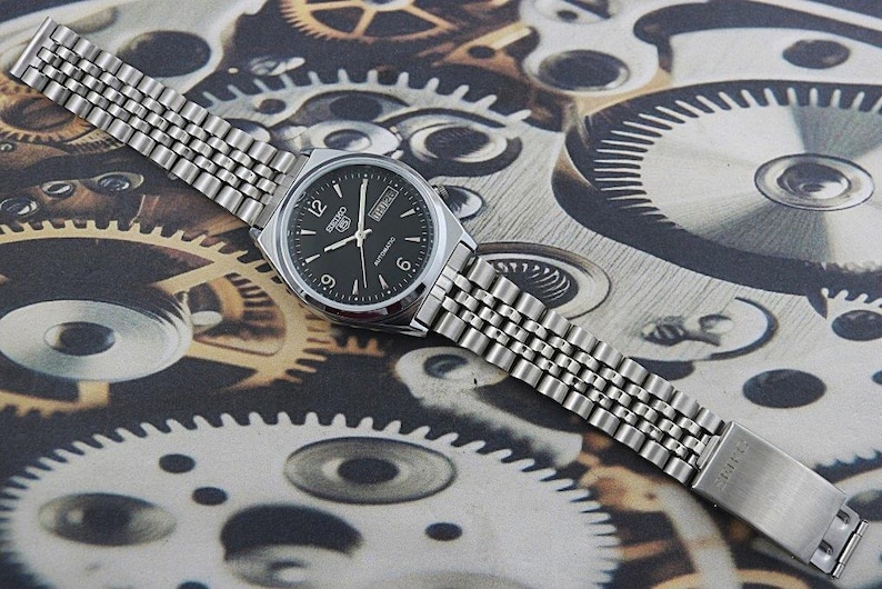 May include: A silver-toned Seiko automatic wristwatch with a black dial, featuring white hour markers and hands. The watch has a stainless steel band and is displayed on a background of gears and mechanical components.