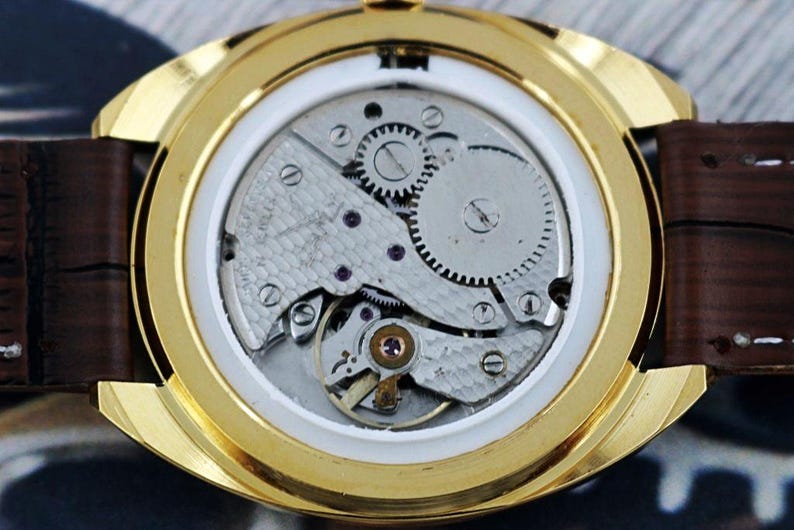 K&ouml;nnte beinhalten: Nahaufnahme des R&uuml;ckens einer goldfarbenen Armbanduhr, die das komplizierte mechanische Uhrwerk zeigt. Die Uhr hat ein braunes Lederarmband und ein wei&szlig;es Zifferblatt. Zahnr&auml;der, Schrauben und andere Komponenten sind sichtbar.