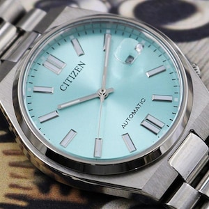 May include: A close-up of a Citizen automatic wristwatch with a light blue face and silver hands and markers. The watch has a silver stainless steel band. The word "Citizen" is printed on the watch face.