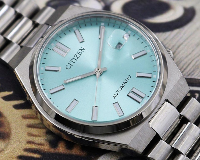 May include: A close-up of a Citizen automatic wristwatch with a light blue face and silver hands and markers. The watch has a silver stainless steel band. The word "Citizen" is printed on the watch face.