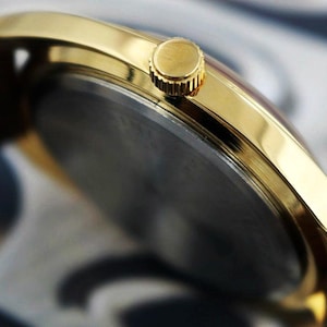 K&ouml;nnte beinhalten: Nahaufnahme einer goldfarbenen Armbanduhr mit braunem Lederarmband. Das Zifferblatt der Uhr ist dunkel, und die Krone ist sichtbar. Die Uhr hat ein klassisches, elegantes Design.
