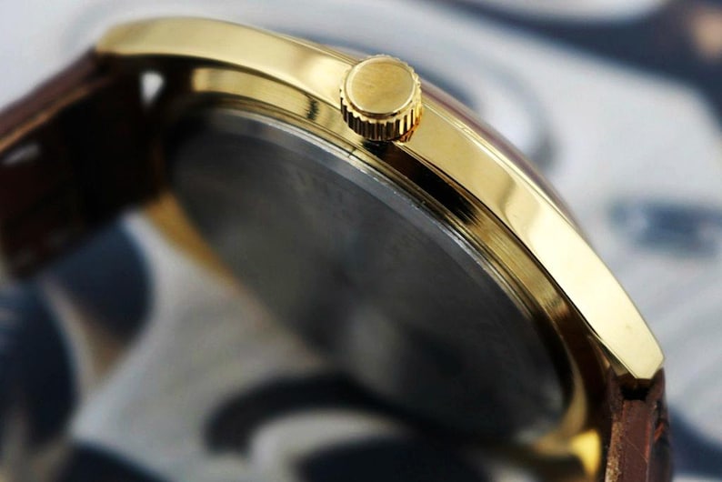 K&ouml;nnte beinhalten: Nahaufnahme einer goldfarbenen Armbanduhr mit braunem Lederarmband. Das Zifferblatt der Uhr ist dunkel, und die Krone ist sichtbar. Die Uhr hat ein klassisches, elegantes Design.