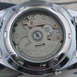 May include: Close-up of the back of a silver wristwatch, showcasing the intricate internal mechanism. The watch face is transparent, revealing gears, springs, and the inscription "JAPAN". The watch band is made of linked silver metal.