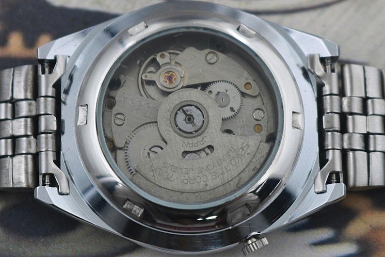 May include: Close-up of the back of a silver wristwatch, showcasing the intricate internal mechanism. The watch face is transparent, revealing gears, springs, and the inscription "JAPAN". The watch band is made of linked silver metal.
