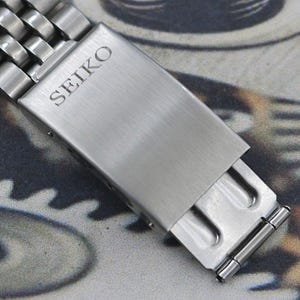 May include: Close-up of a silver-toned watch band clasp. The clasp is rectangular with the brand name "SEIKO" engraved on the top. The band is made of multiple small, connected metal links.