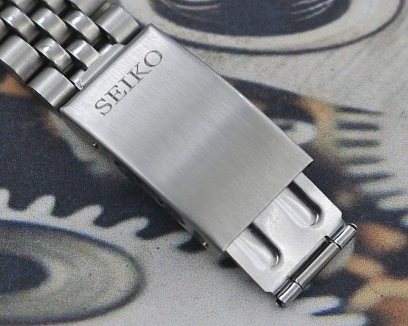 May include: Close-up of a silver-toned watch band clasp. The clasp is rectangular with the brand name "SEIKO" engraved on the top. The band is made of multiple small, connected metal links.