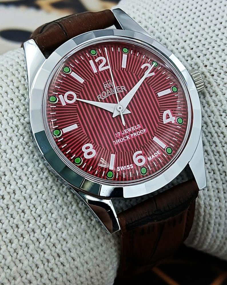 May include: A vintage wristwatch with a red dial and a brown leather strap. The watch has a silver-colored case and white numerals. The dial reads "RW Roamer" and "17 Jewels Shock Proof". The watch is approximately 1.5 inches in diameter.