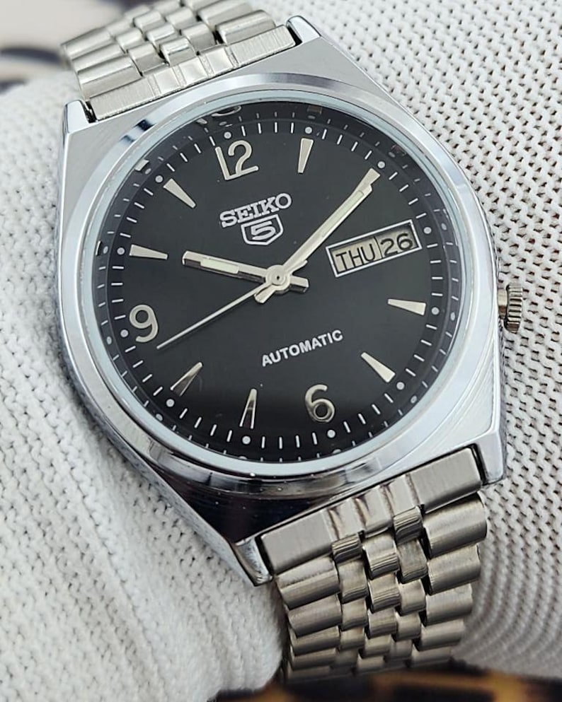 May include: A silver-toned Seiko 5 automatic watch with a black dial. The watch features silver hands and markers, a day-date display, and a stainless steel band. The watch face displays the day as "THU 26".