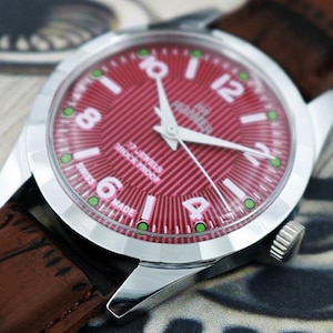 May include: A close-up of a vintage wristwatch with a red dial and white numerals. The watch has a silver case and a brown leather strap. The dial reads "17 Jewels Shock Proof Swiss Made".