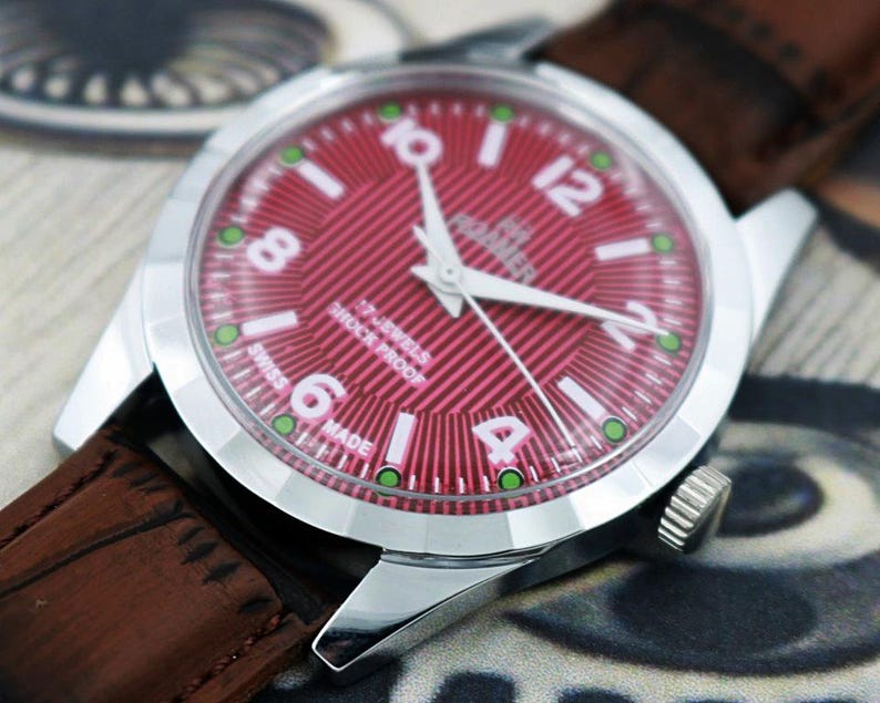May include: A close-up of a vintage wristwatch with a red dial and white numerals. The watch has a silver case and a brown leather strap. The dial reads "17 Jewels Shock Proof Swiss Made".