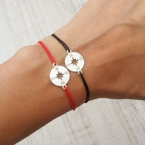 Puede incluir: Dos pulseras de cuerda roja y negra con un dije de plata en forma de brújula. El dije de la brújula tiene una estrella en el centro y las letras N, S, E y W alrededor del borde.