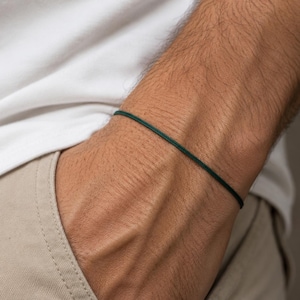 May include: A thin, dark forest green cord bracelet worn around the wrist. The bracelet consists of a single, smooth, round string material that forms a simple, minimalist band encircling the arm without any visible clasps, charms, or metal hardware.