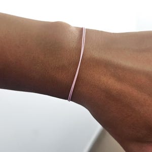 Puede incluir: Una delicada pulsera de hilo rosa claro. El fino cordón está envuelto alrededor de una muñeca, mostrando un diseño minimalista. La pulsera es un accesorio sencillo, perfecto para el uso diario.