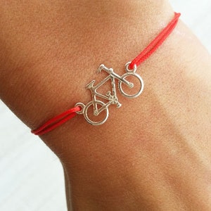 May include: A red string bracelet with a silver bicycle charm. The charm is a detailed silhouette of a bicycle with two wheels.