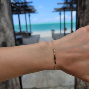 Op de afbeelding: Een delicaat gouden armbandje met een klein kettinkje en een rij kleine vierkante kralen, gedragen op een pols. De achtergrond is een wazige strandscène.