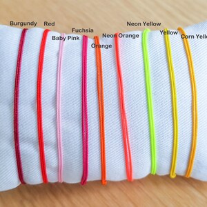 May include: A white pillow with ten different colored cords laid out on it. The cords are labeled with their colors: Burgundy, Red, Baby Pink, Fuchsia, Orange, Neon Orange, Neon Yellow, Yellow, and Corn Yellow.