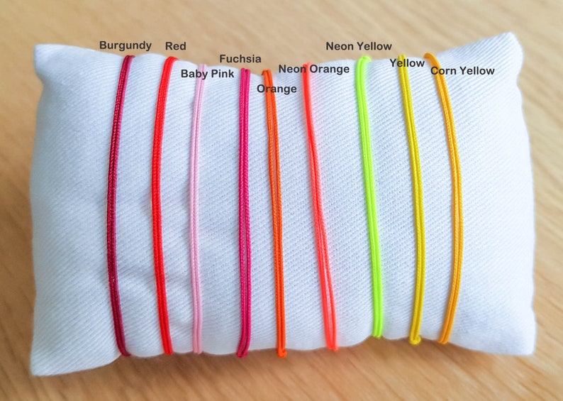 May include: A white pillow with ten different colored cords laid out on it. The cords are labeled with their colors: Burgundy, Red, Baby Pink, Fuchsia, Orange, Neon Orange, Neon Yellow, Yellow, and Corn Yellow.