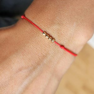 May include: A red string bracelet with five small, gold-colored beads. The bracelet is tied with a knot and is worn on a wrist. The bracelet is a simple, minimalist design.