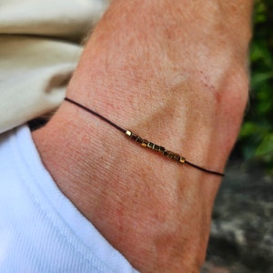 Può includere: Un braccialetto semplice con un cordoncino nero e perline quadrate dorate.