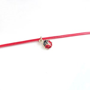 May include: A red string bracelet with a ladybug charm. The charm is red, black, and gold. The bracelet is set against a white backdrop.