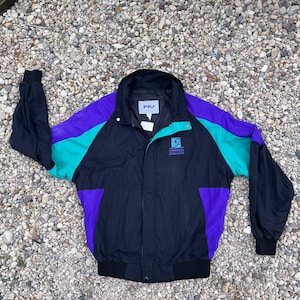 May include: A black, purple, and teal color-blocked windbreaker jacket with a high collar, long sleeves with elastic cuffs, and a front zipper closure. The jacket features a small logo on the left chest and a brand label on the inner neck.
