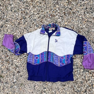 May include: A zip-up windbreaker jacket featuring a color-blocked design in white, deep blue, and purple. The jacket includes a horizontal band across the chest and sleeves with a multi-colored paisley and geometric pattern and a small embroidered logo.