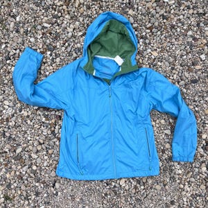 May include: A bright blue zip-up jacket featuring a hood with an olive green interior lining. The jacket includes a full-length front zipper, long sleeves with elasticized cuffs, and two vertical zippered pockets on the front torso area.