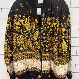 May include: Black bomber jacket with a gold floral pattern and a white and gold border. The jacket has a black ribbed collar, cuffs, and hem, and a full-length zipper. The jacket is hanging on a black hanger against a white brick wall.