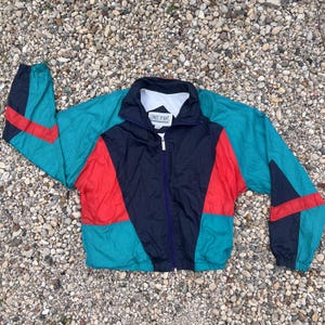 May include: A color-blocked windbreaker jacket featuring a navy blue center panel, teal sleeves and lower sections, and bright red accents. The jacket has a full-length front zipper, a high collar with a white inner lining, and a Sands Point brand label.