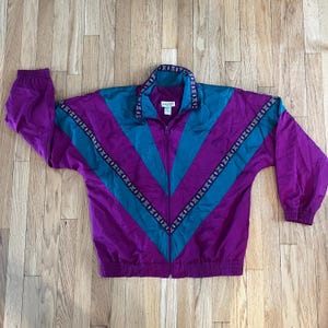 May include: A vintage purple and teal zip-up track jacket with a chevron design. The jacket features a high collar and decorative trim along the collar and sleeves. The jacket is made of a shiny, lightweight material.