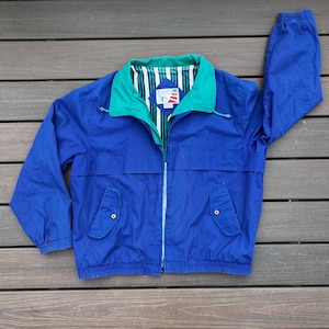 May include: A blue, green, and white windbreaker jacket with a zipper closure. The jacket has a green collar and a white and green striped lining. The jacket has two pockets on the front.