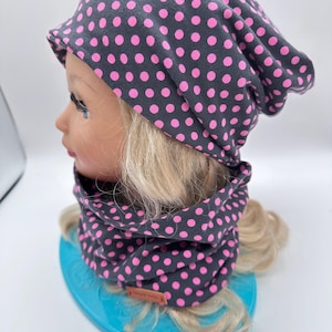 Könnte beinhalten: Ein dunkelgraues Mütze- und Halstuch-Set mit rosa Tupfenmuster. Die Mütze ist locker geschnitten, und das Halstuch ist röhrenförmig. Das Set wird auf einem Schaufensterkopf mit blonden Haaren präsentiert.