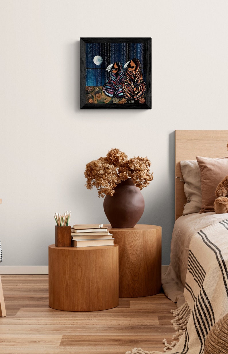 Puede incluir: Una obra de arte enmarcada que representa a dos figuras con ropa colorida sentadas en una superficie con patrones, con una luna llena en el fondo.