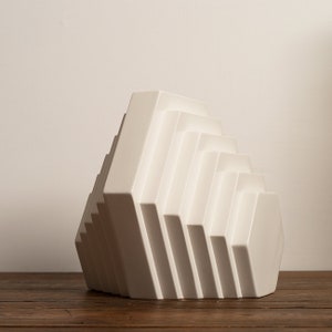 May include: White ceramic geometric vase with a layered, stepped design. The vase is shaped like a pyramid with a hexagonal base.