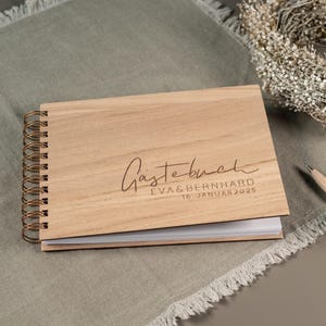 May include: A wooden guest book with a gold spiral binding. The cover is engraved with the text "G&auml;stebuch" and the names "Eva & Bernhard" and the date "16. Januar 2025".