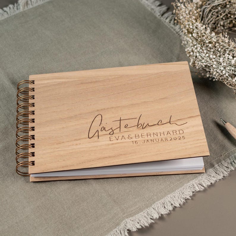 May include: A wooden guest book with a gold spiral binding. The cover is engraved with the text "G&auml;stebuch" and the names "Eva & Bernhard" and the date "16. Januar 2025".