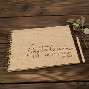 May include: A wooden guest book with a spiral binding. The cover is engraved with the text "G&auml;stebuch" in cursive, followed by "Eva-Maria & Markus" and "15. Juli 2023".