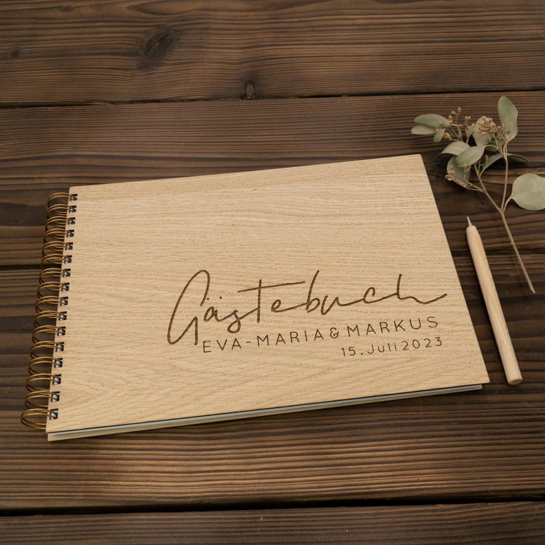 May include: A wooden guest book with a spiral binding. The cover is engraved with the text "G&auml;stebuch" in cursive, followed by "Eva-Maria & Markus" and "15. Juli 2023".