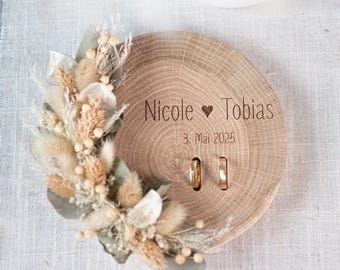 May include: Wooden ring holder with a dried floral arrangement and two gold rings. The holder is engraved with "Nicole ♥ Tobias" and "3. Mai 2025".
