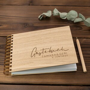 May include: A wooden guest book with a gold spiral binding. The cover is engraved with the text "G&auml;stebuch TAMARA & GERIC 9. JUNI 2023".