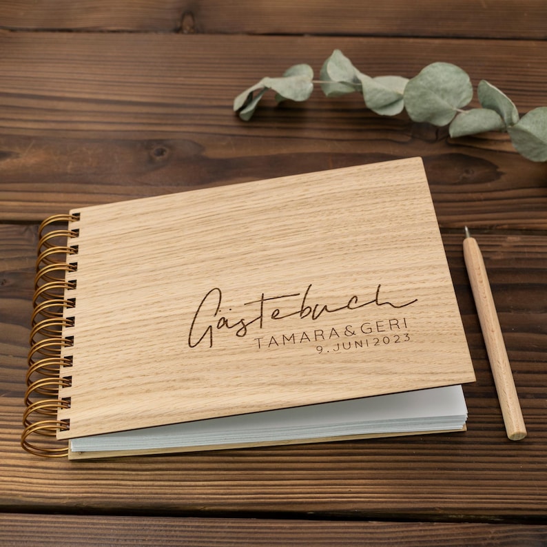 May include: A wooden guest book with a gold spiral binding. The cover is engraved with the text "G&auml;stebuch TAMARA & GERIC 9. JUNI 2023".