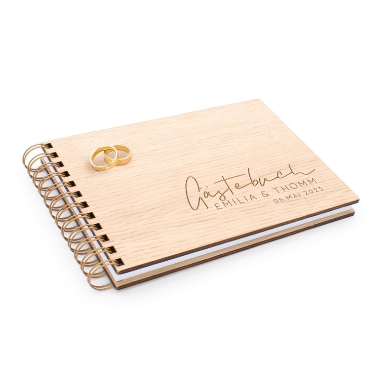 May include: A wooden guest book with a spiral binding. The cover is light brown and has the text "G&auml;stebuch" in gold lettering, followed by "Emilia & Thomm 06. Mai 2023". Two gold rings are placed on the cover.