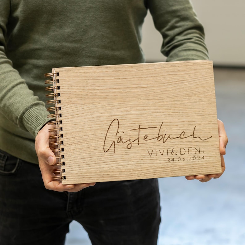 May include: A wooden guest book with a spiral binding. The cover is engraved with the text "G&auml;stebuch" in cursive script, followed by "Vivi & Deni" and the date "24.05.2024".