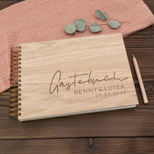 May include: A wooden guest book with a spiral binding. The cover is engraved with the text "G&auml;stebuch" and the names "Benny & Luisa" and the date "20.05.2023".