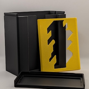 May include: A black rectangular storage box with a separate black lid and a yellow front panel featuring a jagged, vertical cutout design. The box has a textured finish and is constructed from rigid material, showing a modular assembly.