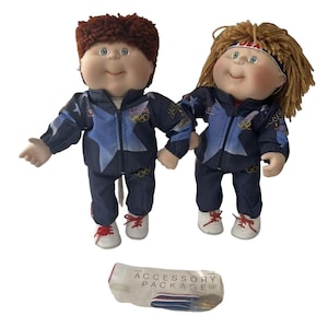 May include: Two Cabbage Patch Kids dolls wearing blue and white tracksuits with the 1996 Olympic logo. The dolls have red and blue accents on their outfits and are holding a small white package that says "Accessory Package".