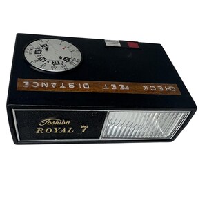 May include: A black Toshiba Royal 7 camera with a circular dial and a rectangular flash. The dial displays numbers and measurements in feet and meters. The words "CHECK DISTANCE FEET" are printed on a wooden strip.