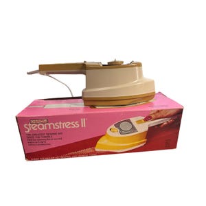 May include: A vintage Osrow Steamstress II sewing aid, featuring a cream and tan iron with a pink box. The box text reads "The Greatest Sewing Aid Since The Thimble". The iron is designed for opening seams and darts.
